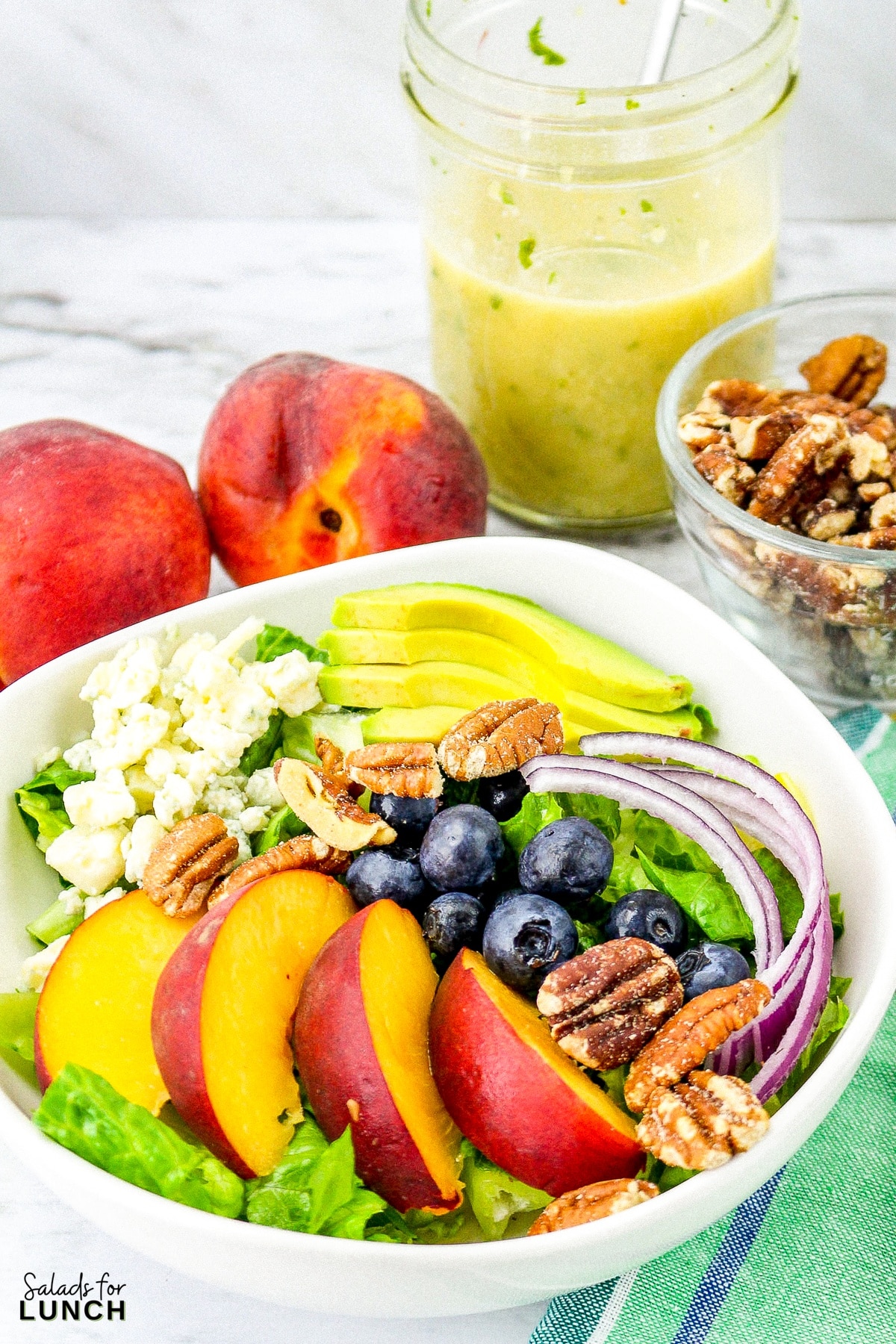 Summer peach salad with fresh peaches, blueberries, onion, romaine lettuce, pecans and blue cheese in a square white bowl with fresh mint dressing in the background on a green napkin and a white marble surface