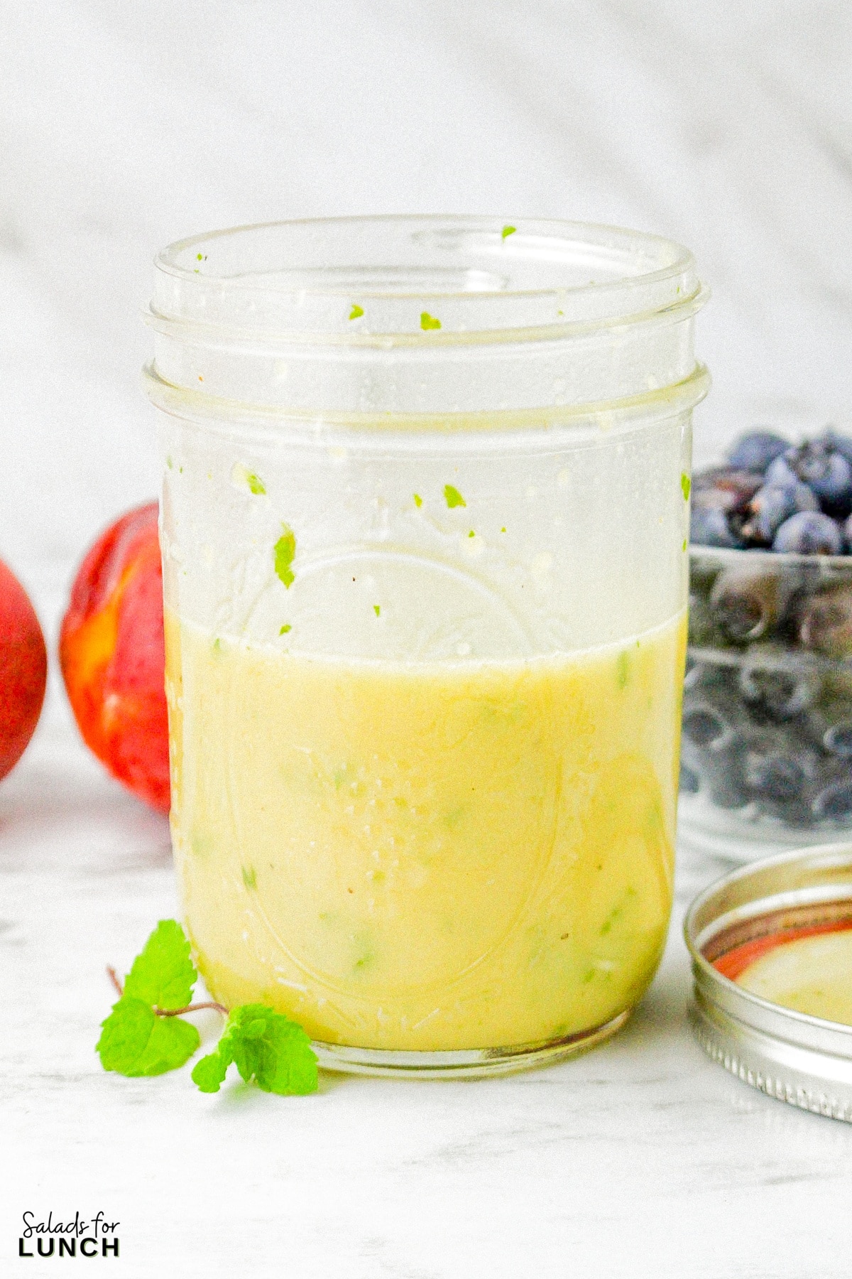 Fresh mint salad dressing in a mason jar with a sprig of fresh lint beside the jar, and a bowl of fresh blueberries, and two peaches in the background