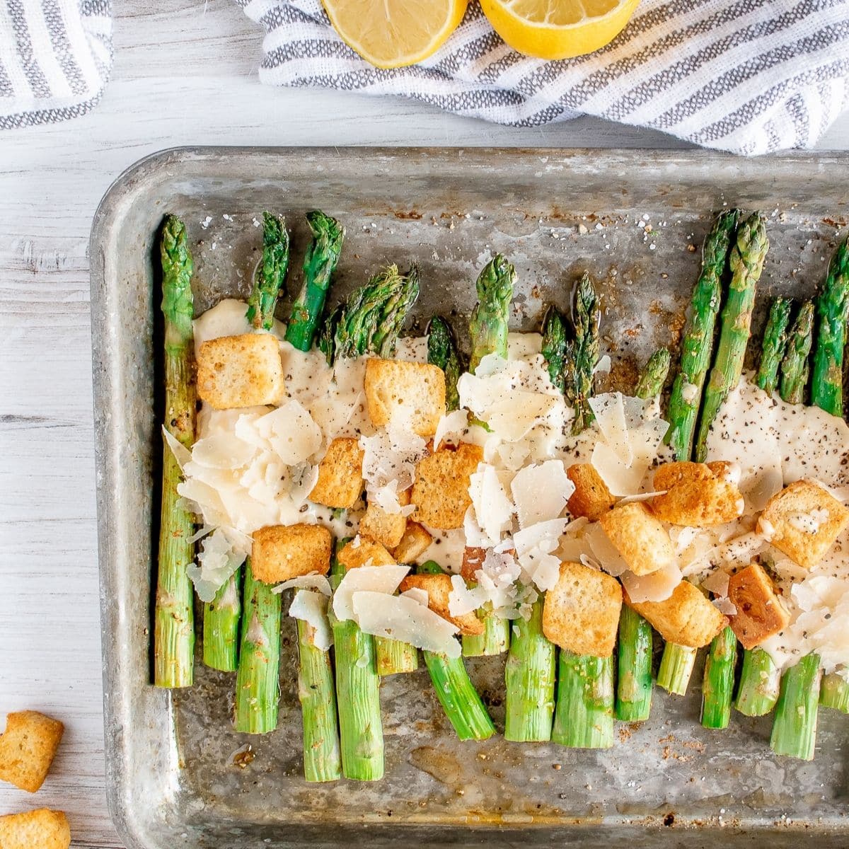 Roasted asparagus lined in a row on a baking sheet drizzled with creamy caesar salad dressing, fresh parmesan cheese and crispy croutons.