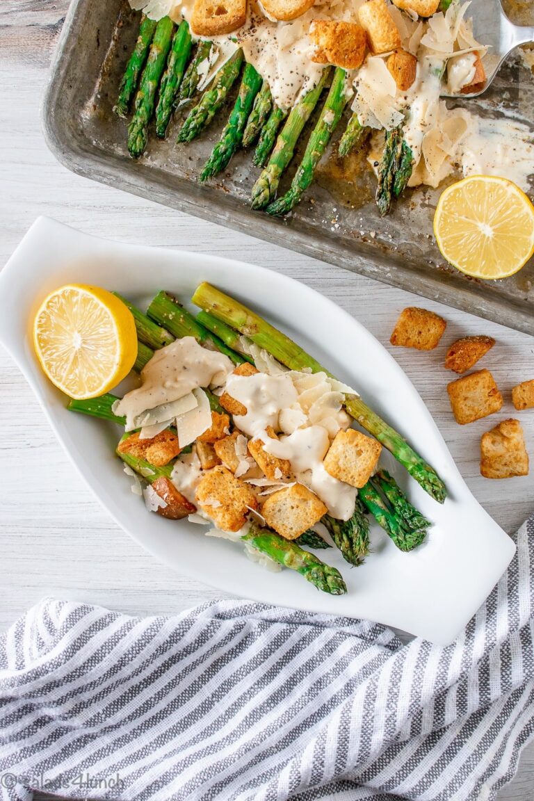 Roasted asparagus lined in a row in an oval white bowl drizzled with creamy caesar salad dressing, fresh parmesan cheese and crispy croutons.