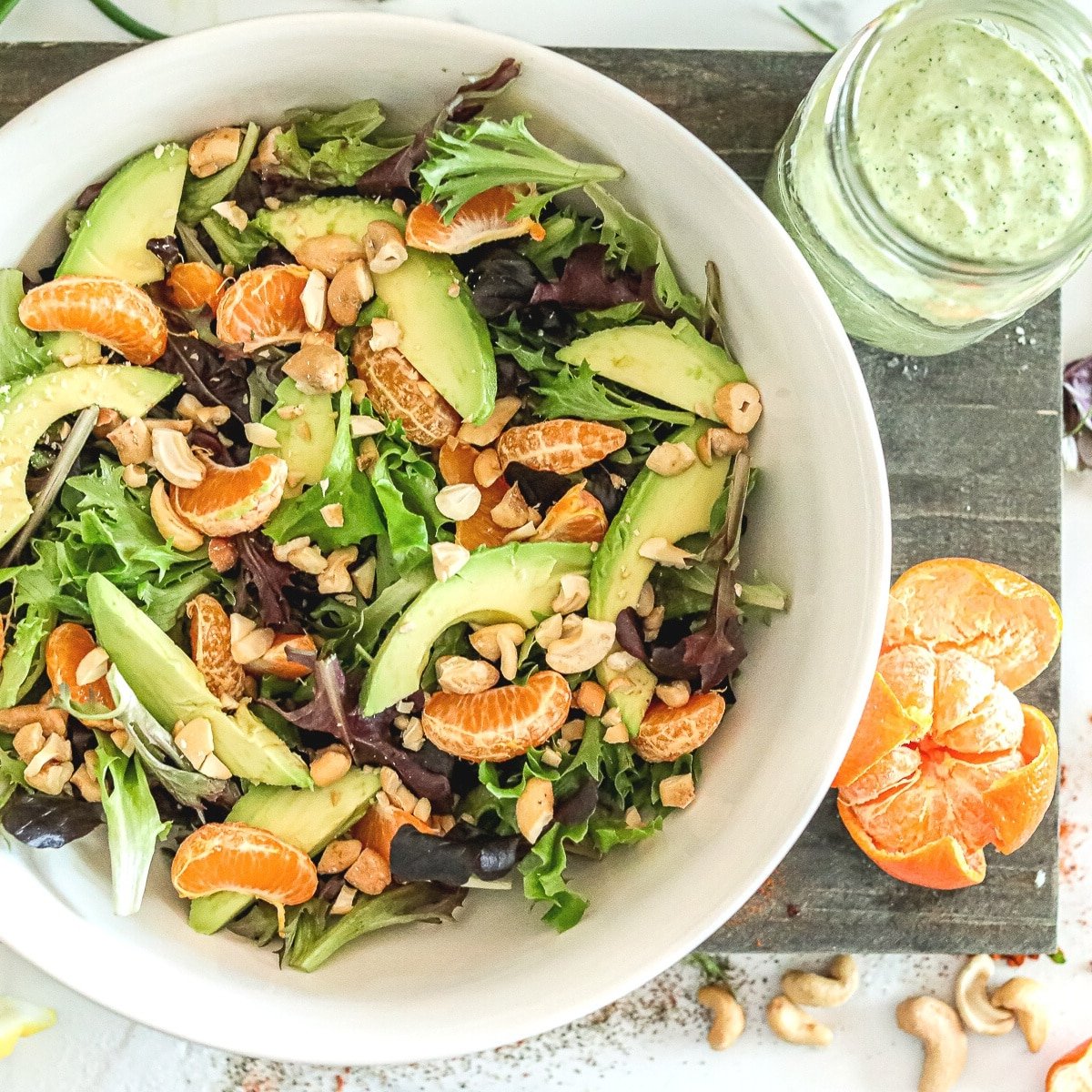 a white bowl with spring salad, mandarin oranges and cashews on a cutting board besid a mason jar with green goddess salad dressing.
