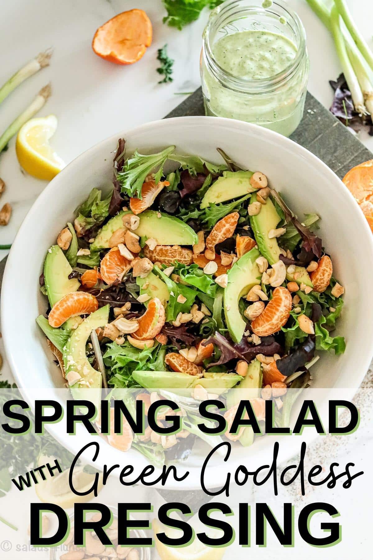 A white bowl with spring salad, mandarin oranges and cashews on a cutting board besid a mason jar with green goddess salad dressing.