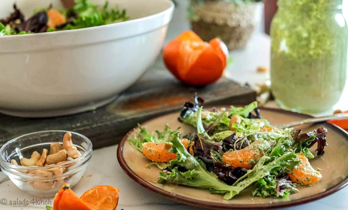 Close up of a brown plate with a colourful spring salad with mandarin oranges and small bowl of cashews, and a large jar of green goddess dressing beside it.