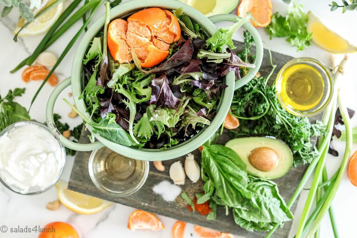 Overhead photo of the ingredients needed to make a spring salad with green goddess dressing: spring salad mix, mandarin oranges, cashews, avocado, plain greek yogurt, olive oil, apple cider vinegar, lemon juice,fresh basil, fresh parsley, and green onion.