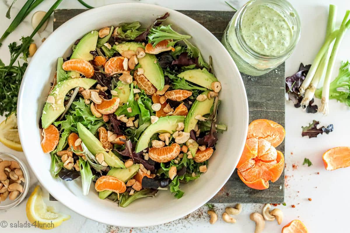 A white bowl with spring salad, mandarin oranges and cashews on a cutting board beside a mason jar with green goddess salad dressing.