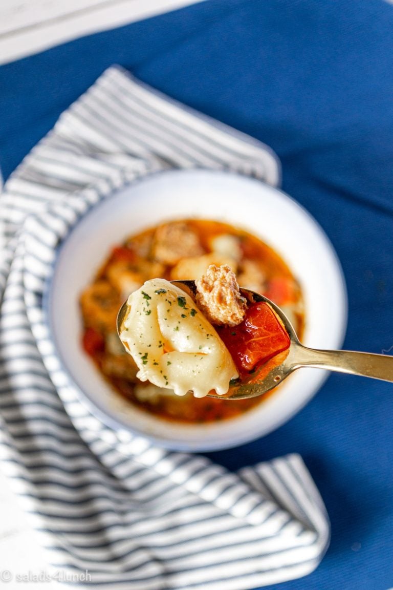 Close up of italian sausage and tortellini soup on a spoon with a white bowl of soup on a blue and white striped napkin in the background.