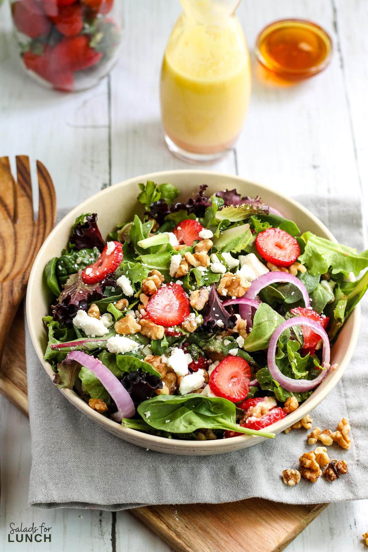 Overhead photo of a white bowl with a colourful strawberry, feta and mint salad with spring mix on a grey napkin.