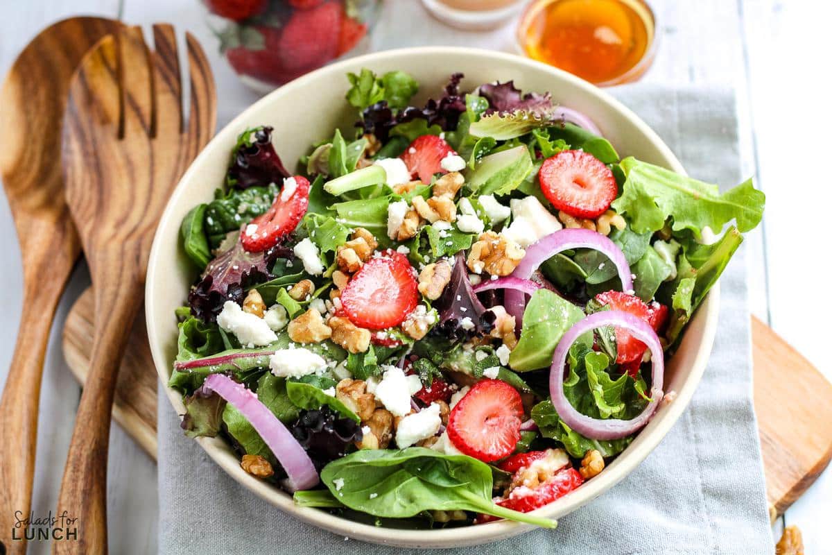 White bowl of a colourful strawberry, feta and mint salad with spring mix on a grey napkin with wooden salad spoons beside the bowl.