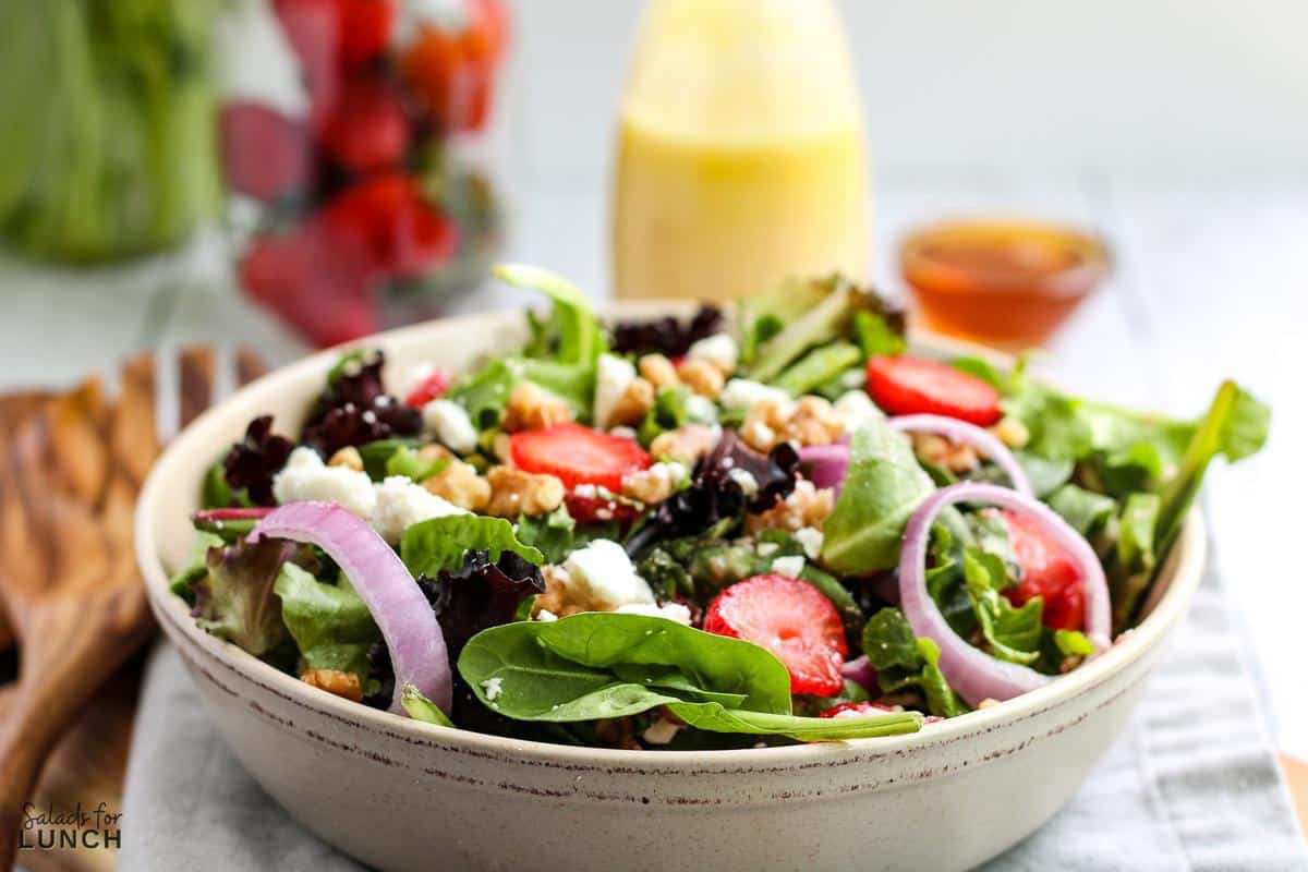Strawberry feta and mint salad with fresh greens in a white bowl with honey dressing and strawberries in the background.