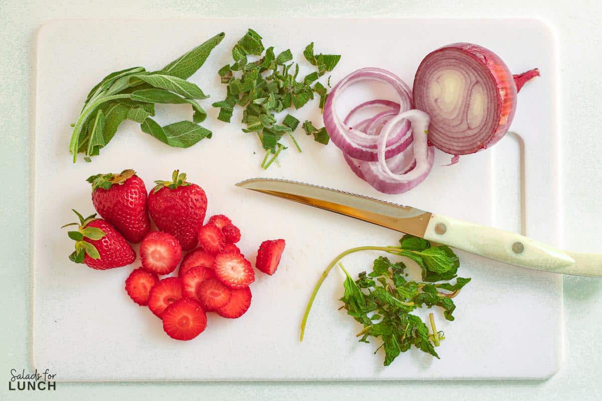 White cutting board with white knife and sliced strawberries, red onion, mint and sage leaves.