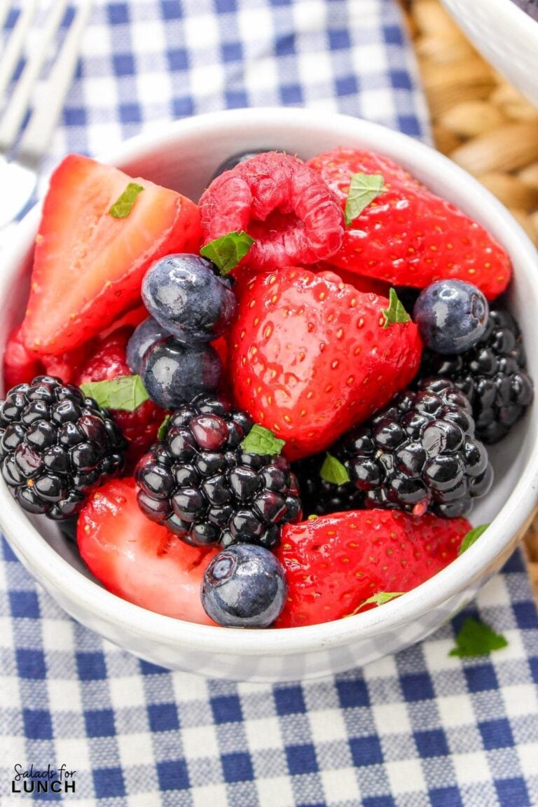 White bowl with summer berry salad featuring sliced strawberries, blue berries and black berries on a blue and white checkered napkin.