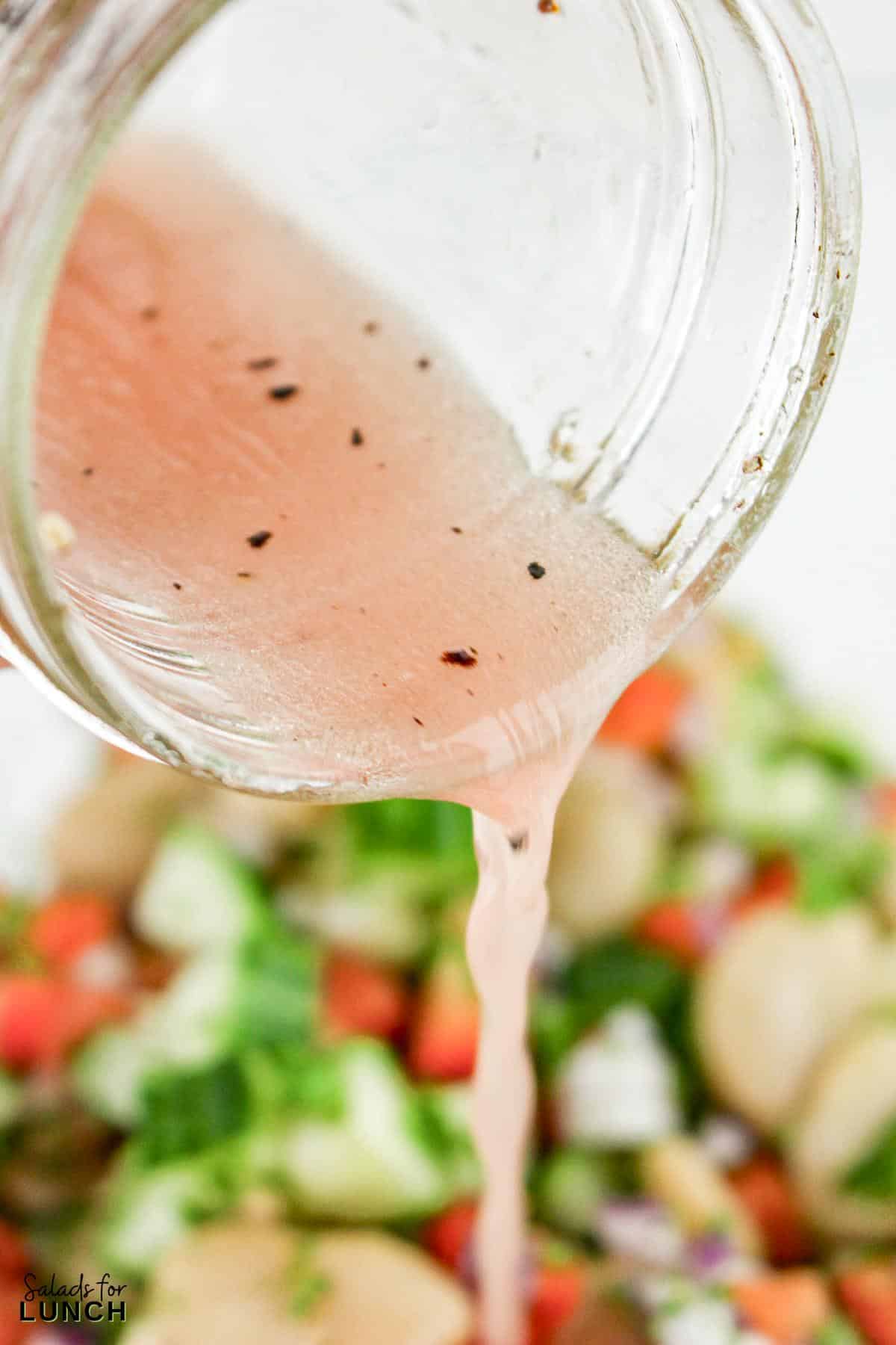 A glass jar pouring pink red wine vinegar dressing onto a salad.