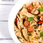 White bowl of bow tie feta bruschetta pasta salad with sprig of basil and roma tomatoes.